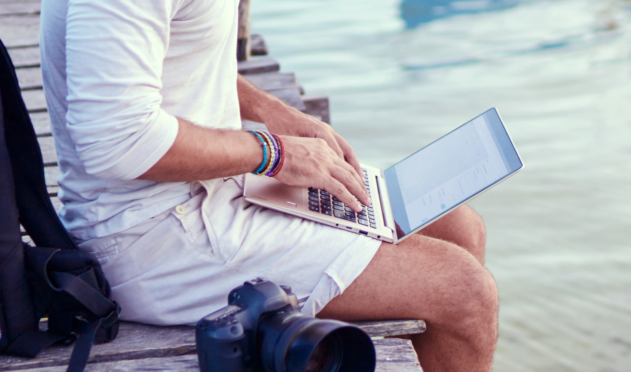 A person is working on their laptop from a park bench. Located next to them is their DSLR camera.